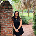 Gabi is pictured in a black dress leaning against a brick column. She has dark hair, fair skin, and is smiling.