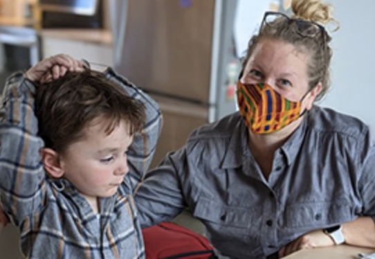 Dr. Caitlin Hudac, a White woman with curly blonde hair in a bun, wears a mask and poses next to a young, White boy putting his arms over his head.