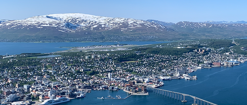 Panoramic aerial view of Tromsø, Norway, showing the coastal city, harbor, bridge and surrounding snow-capped mountains under a clear blue sky.