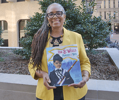 A smiling woman with long gray dreadlocks and glasses stands outdoors in front of a building with trees and shrubs. She wears a yellow blazer over a black and gold patterned top and holds a children's book titled 'Ida B. Wells Marches for the Vote.' The book cover features an illustration of Ida B. Wells, a young Black woman with dark hair in an updo, wearing a black and white dress with a bowtie, holding a protest banner against a background of marchers. A close-up of the book cover is also shown on the right side of the image.