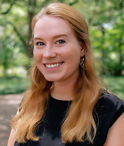 A young woman with long, wavy blonde hair smiles at the camera. She is wearing a black sleeveless top and gold, geometric-shaped earrings. The background is blurred with lush green trees and foliage, suggesting an outdoor setting