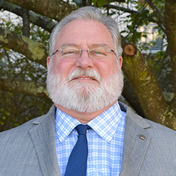 a man with a gray beard and hair wearing glasses, with a blue tie and gray jacked on standing outside