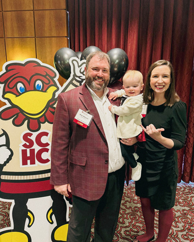 Smiling couple holding their young daughter while standing next to a cardboard cutout of Cocky.