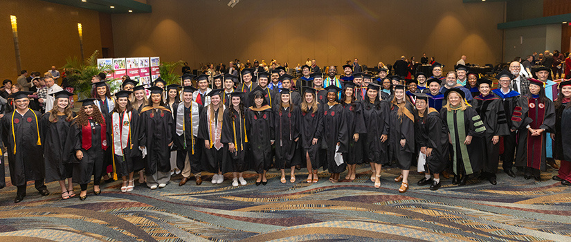 A large group of HRSM graduates in black caps and gowns, along with faculty members in academic regalia, pose for a group photo in a spacious indoor venue. Some graduates wear honor cords and stoles, and the faculty members are dressed in colorful robes and hoods. The background includes additional attendees and informational displays.