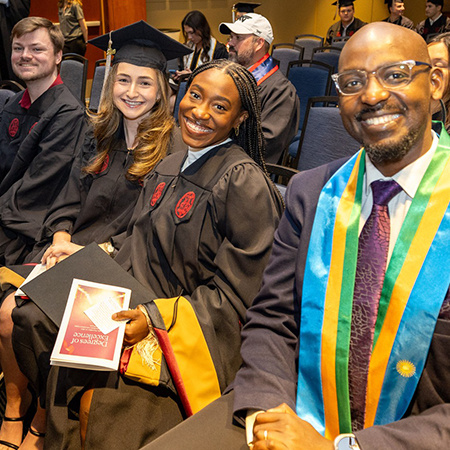 A group of graduates from the College of Hospitality, Retail and Sport Management smile as they sit in their caps and gowns at the Degrees of Excellence celebration.