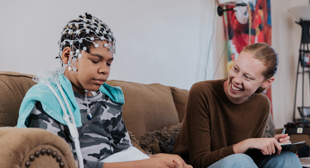 Wearing an EEG net on his head, AJ Peterman sits on his couch beside research assistant Ashlan Cheever, who is engaging him in conversation.